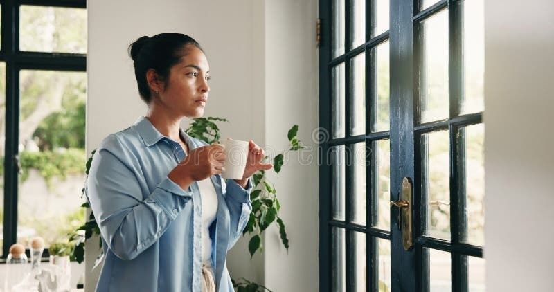 Frau mit Tasse steht nachdenklich an einem großen Fenster